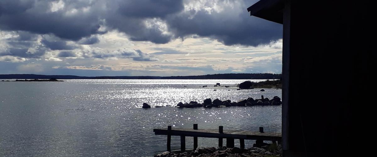 Färgfoto på sjöutsikt från stranden. I förgrunden finns knuten av en båthus och en kaj. Solen glittrar på vattenytan men på skyn vandrar tunga moln.