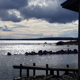 Färgfoto på sjöutsikt från stranden. I förgrunden finns knuten av en båthus och en kaj. Solen glittrar på vattenytan men på skyn vandrar tunga moln. 