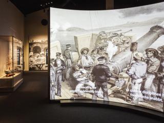 Interiör Bomarsunds besökscenter. I förgrunden en bildskärm som visar en film, med en litografi i blickfånget. I bakgrunden, till höger om bildskärmen, skymtar ett utställningsrum med en glasmonter.