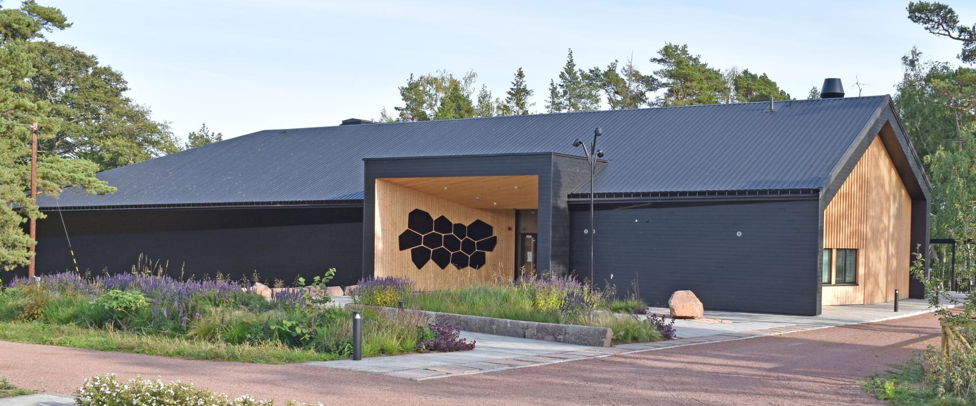 En avlång byggnad med svarta väggar och tak, infälld entré och gavlar i gult trä. Runt om vägar och rabatter.