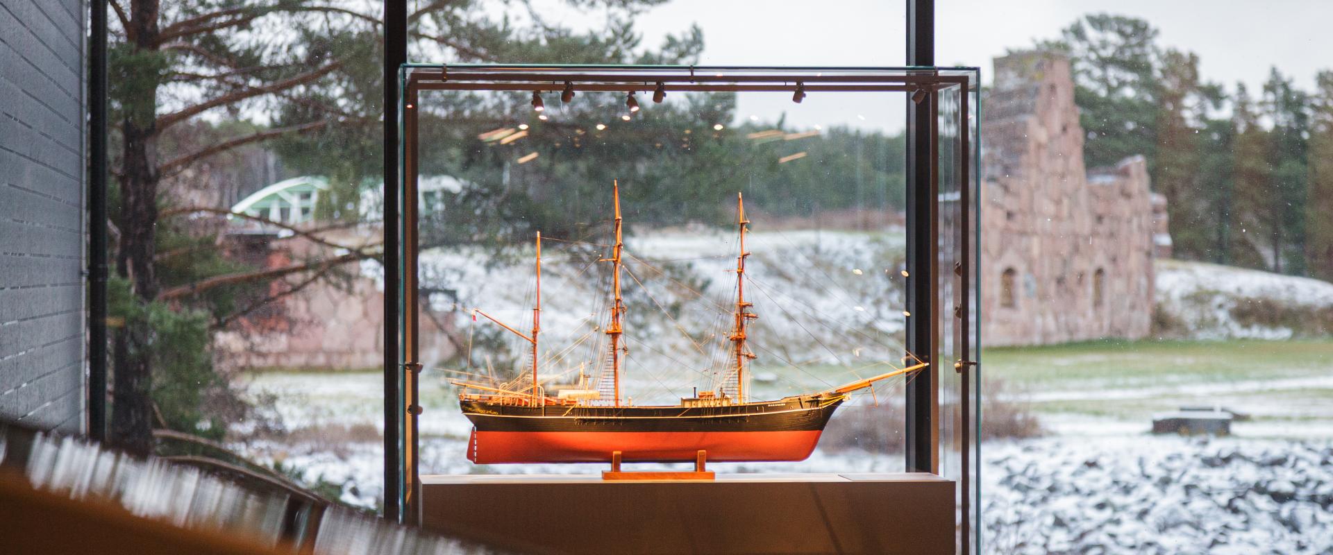Ett modellfartyg i förgrunden. Genom ett fönster ses ett vinterlandskap och ruinerna av en mur, Bomarsunds fästning.