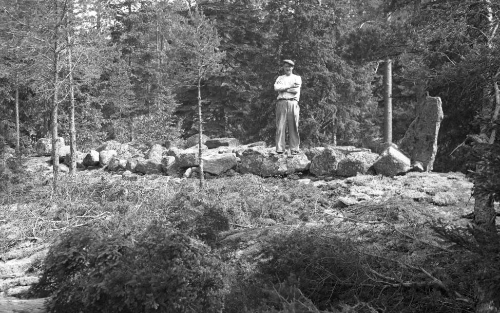Syntolkning: Ett gammalt fotografi i svartvitt. Bilden visar en skeppsformig grav av stenar som är placerad på en bergsklack omgiven av små tallar och större granar. I den högra ändan av skeppet ses ett större rest stenblock. På stenskeppet står en man med armarna i kors. Han bär tidsenliga kläder från 1930-talet med en skärmmössa på huvudet, ljus skjorta och ljusa lössittande byxor. Mannen tittar mot kameran men hans ansikte skuggas av skämmössan.