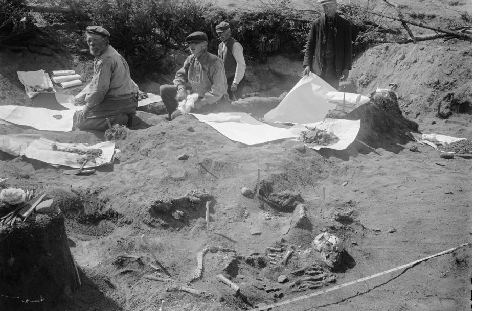 Syntolkning: Ett gammalt fotografi i svartvitt taget i början av 1900-talet. Bilden visar en arkeologisk utgrävning av Jettböle stenåldersboplats. Utgrävningen har skapat större en grop i sanden som är cirka 1 meter djup. Fyra personer sitter och står i gropen, varav tre är äldre män och en är en yngre pojke. Alla bär mössor med skärm framtill och har smutsiga kläder. De arbetar som grovgrävare. På sanden ligger flera vita pappersark och grävverktyg. Närmast i bild har de grävt fram en människoskelett. Skelettet ligger visar att personen har legat på sidan med uppdragna knän. Bredvid skelettet är ett måttband utplacerat.