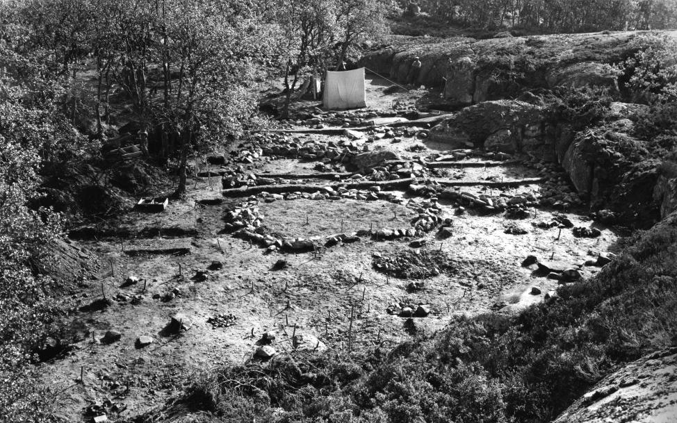 Syntolkning: Ett gammalt fotografi i svartvitt taget från en bergsklack. Bilden visar en platt markyta i mitten som är omgiven av berg på höger sida och en skogsdunge med lövbeklädda träd på vänster sida. På markplätten finns flera större runda cirklar skapade av stenar och är hyddbottnar. I stencirklarna, men även på sidan om, finns små högar av stenar som är kokgropar och eldhärdar. Bak i bilden, bakom hyddbottnarna finns ett enkelt rest tält av textilduk uppsatt mellan träden. Bredvid ses en person som lutar sig mot bergsväggen.