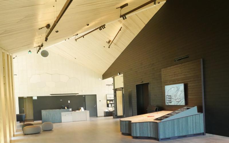 Bild av entréhallen i Bomarsunds besökscenter. Mörk vägg med modell av fästningen till höger, ljus vägg rakt fram och ljust tak. Receptionsdisk rakt fram.