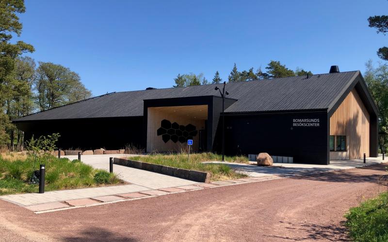 En låg, avlång byggnad med svart tak och väggar, gavel och entré träfärgade. Framför huset finns en grusväg, gångväg och planteringar.